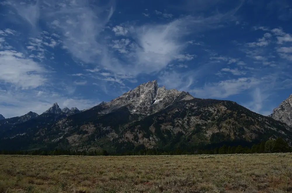 Grand Teton: der höchste Gipfel der Teton Range über dunklen Waldhängen und einer Salbeiebene, Federwolken am Himmel