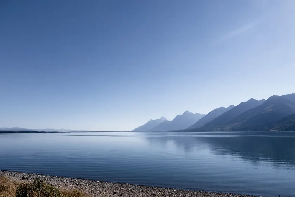Ruhiger Jackson Lake im blauen Abendlicht, am Ufer Kiesel, dahinter die Bergkette der Teton Range
