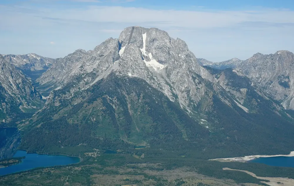 Luftaufnahme des Mount Moran von Osten: massiger Granitgipfel mit Schneefeldern über bewaldeten Hängen und einem See