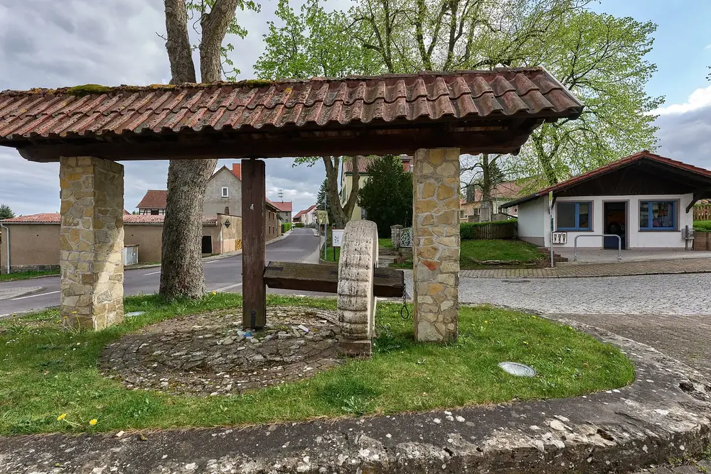 Platz Am Waldstein in Craula: überdachter Brunnen aus Natursteinsäulen, dahinter Dorfhäuser und Bäume