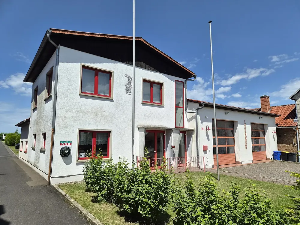 Feuerwehrhaus in Kammerforst: weißes Gebäude mit roten Fenstern und Toren an einer Dorfstraße unter blauem Himmel