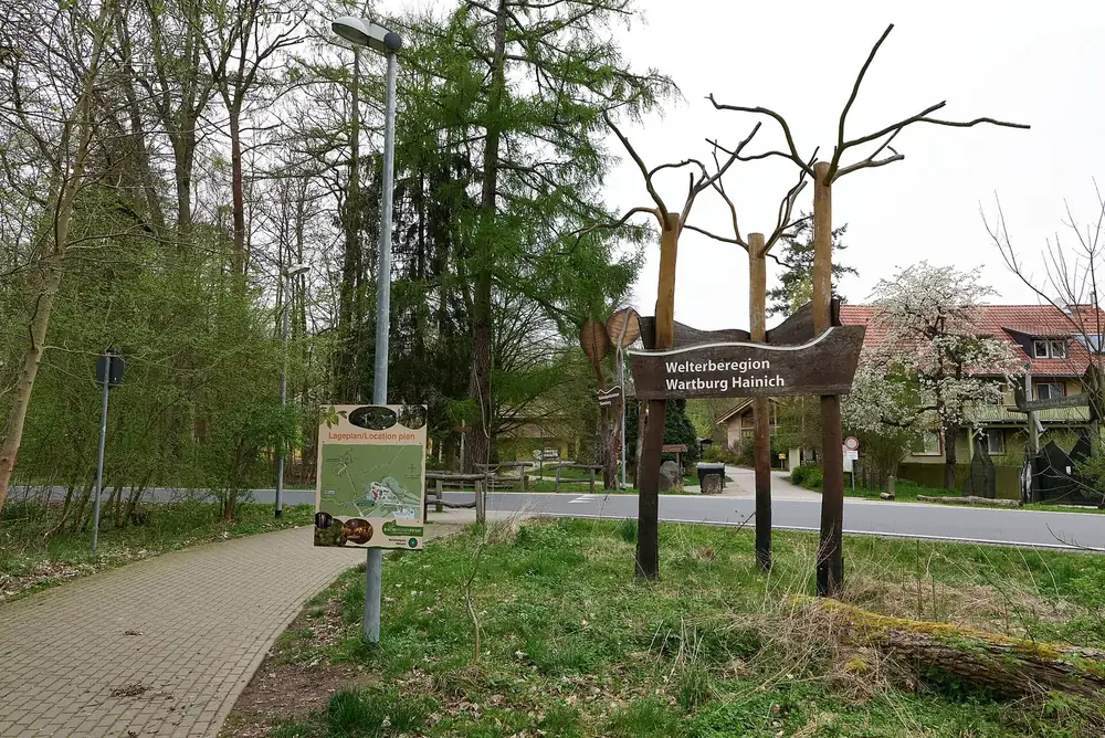 Eingangsbereich an der Thiemsburg im Hainich: gepflasterter Weg, Holzskulptur mit Schild und Infotafeln unter Bäumen