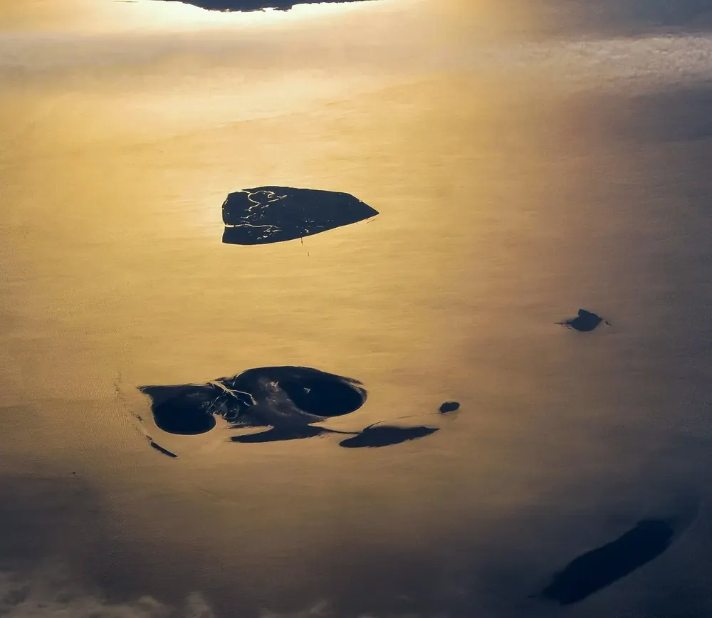 Luftaufnahme im Gegenlicht: Neuwerk, Scharhörn und Nigehörn als dunkle Flächen im goldglänzenden Wattenmeer