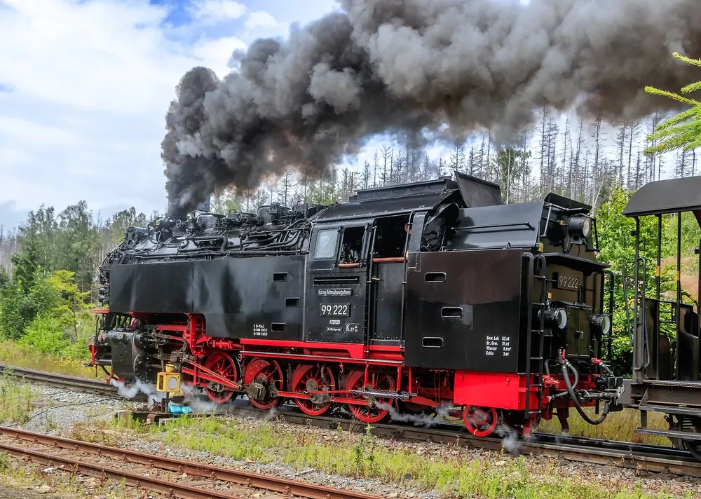 Dampflok 99 222 der Brockenbahn mit schwarzer Rauchwolke im Bahnhof Schierke, dahinter lichter Wald
