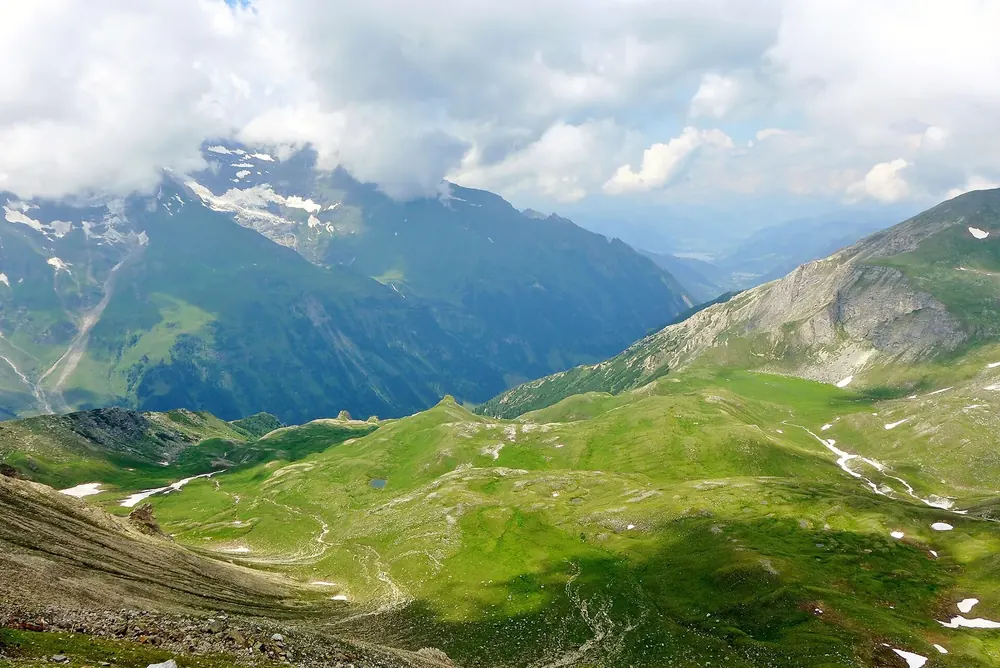 Blick vom Aussichtsturm der Edelweißspitze hinab in grüne Almtäler und auf wolkenverhangene Gipfel der Hohen Tauern