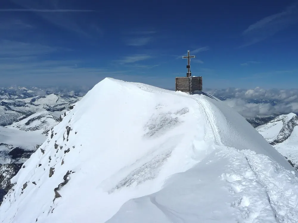 Gipfel des Großvenediger: verschneiter Firngrat mit Gipfelkreuz über einem Meer vergletscherter Berge