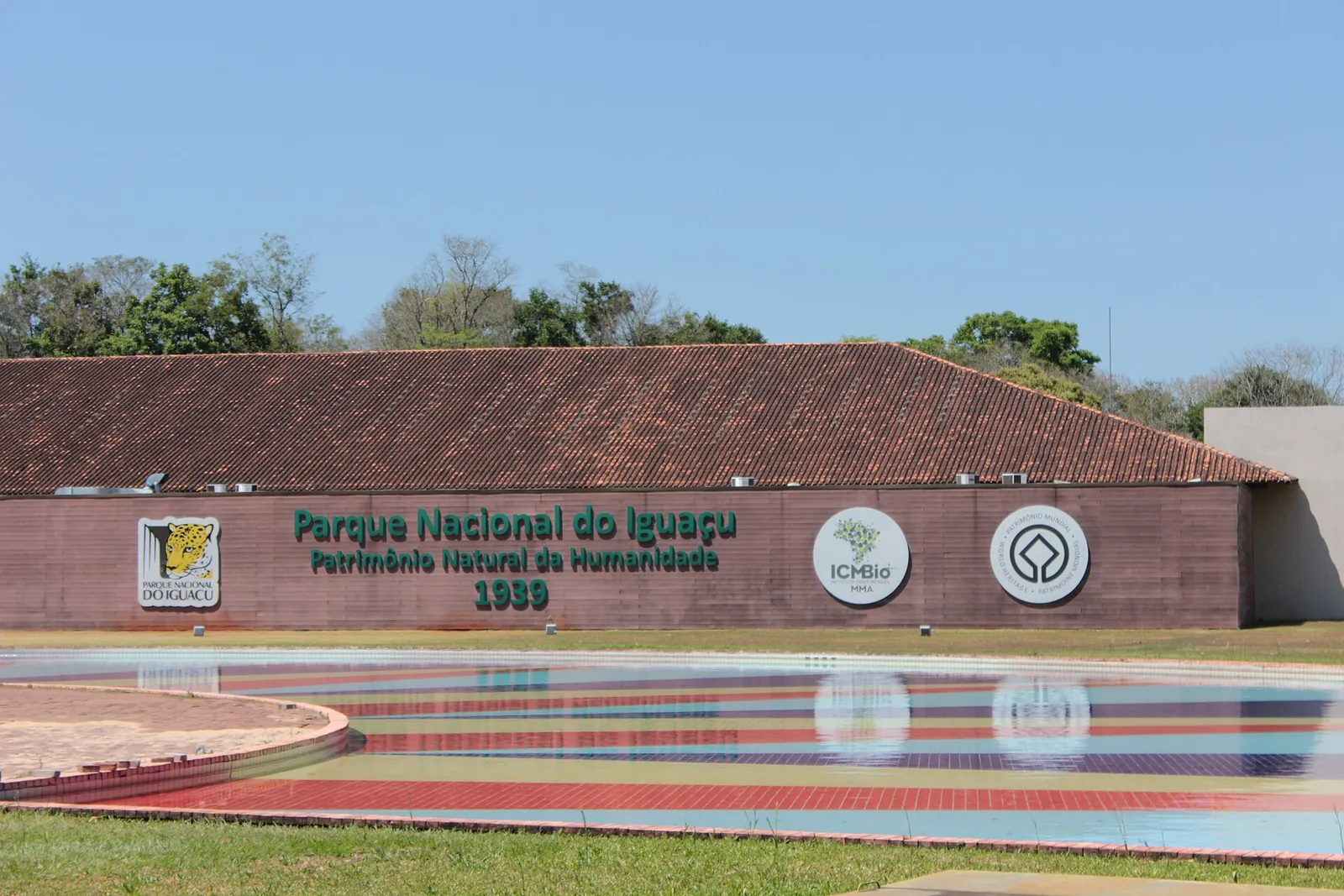 Eingangsgebäude des Iguaçu-Nationalparks mit Ziegeldach und dem Schriftzug „Parque Nacional do Iguaçu – Patrimônio Natural da Humanidade 1939“, davor ein farbiges Wasserbecken