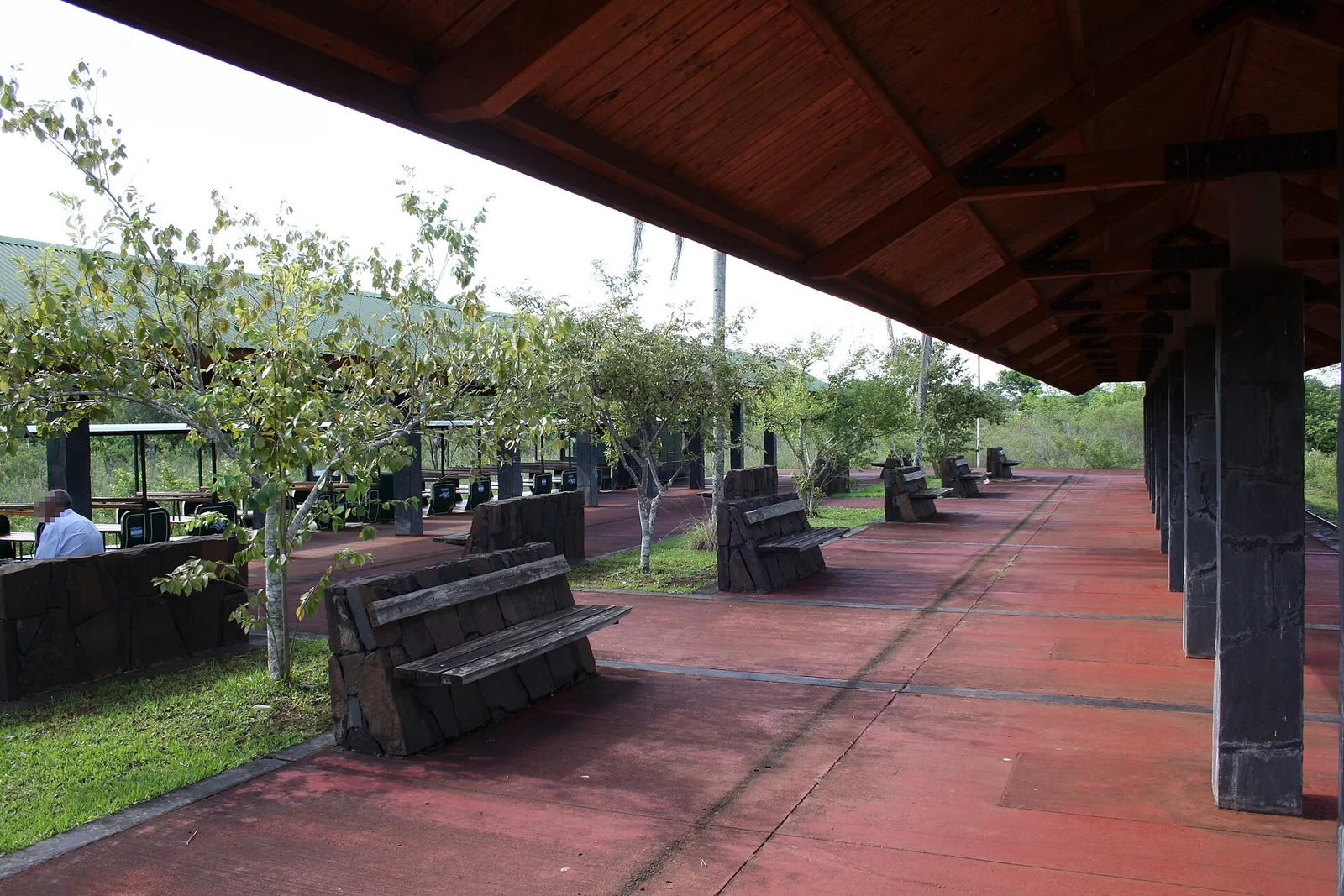 Überdachter Bahnsteig der Estación Central im Iguazú-Nationalpark mit roten Bodenplatten, Holzbänken und Holzdach, daneben Bäume und Wiese