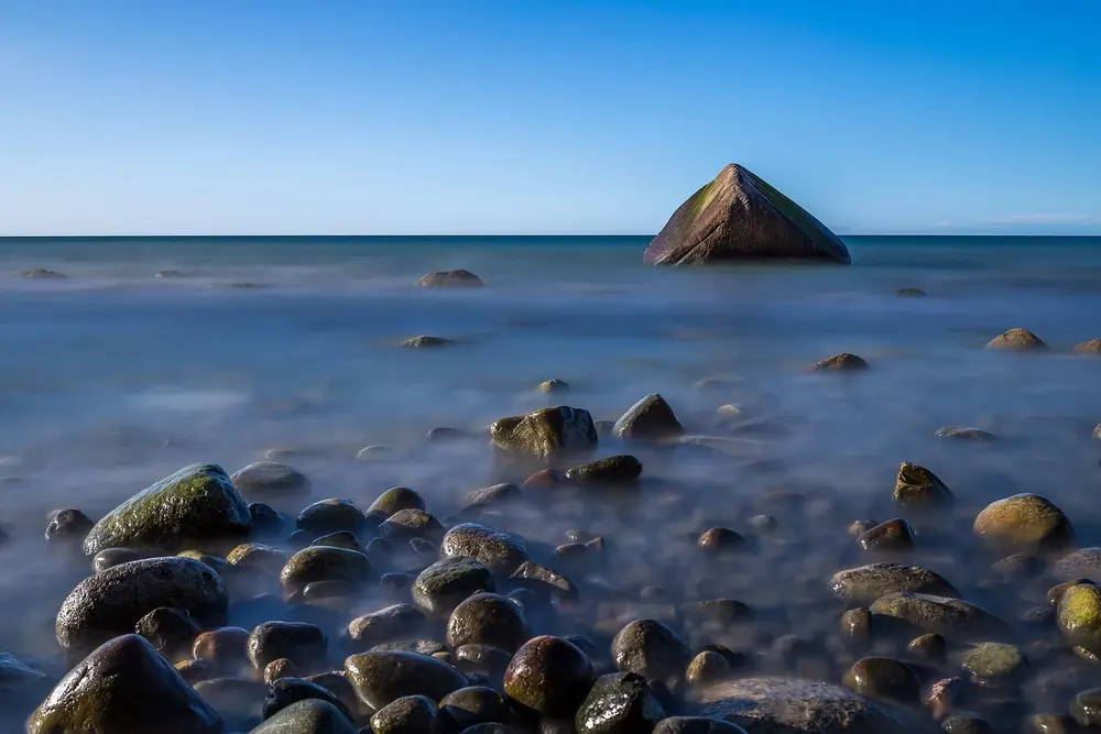 Der Schwanenstein, ein Findling in der Ostsee vor Lohme, davor runde Steine im glatt verwischten Wasser
