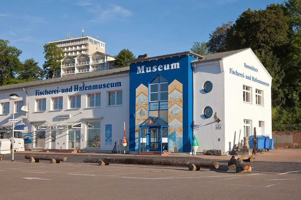 Fischerei- und Hafenmuseum in Sassnitz: weißes Gebäude mit blauem Eingangsbau am Hafen, dahinter ein Hotelhochhaus