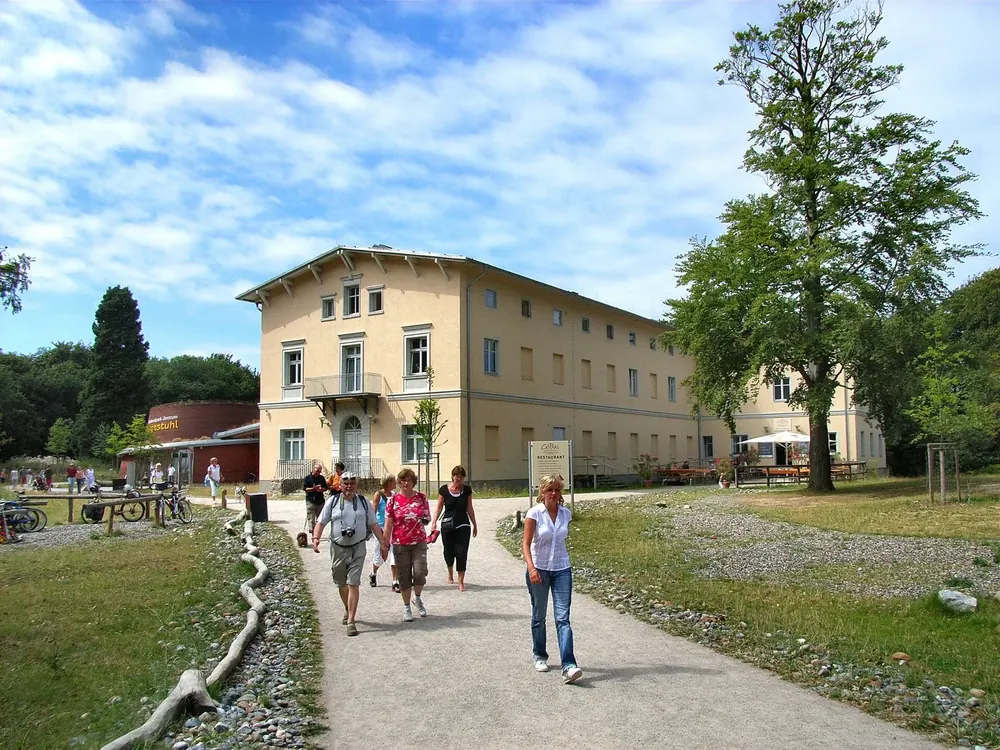 Nationalpark-Zentrum Königsstuhl: helles historisches Gebäude zwischen alten Buchen, davor ein Weg mit Besuchern
