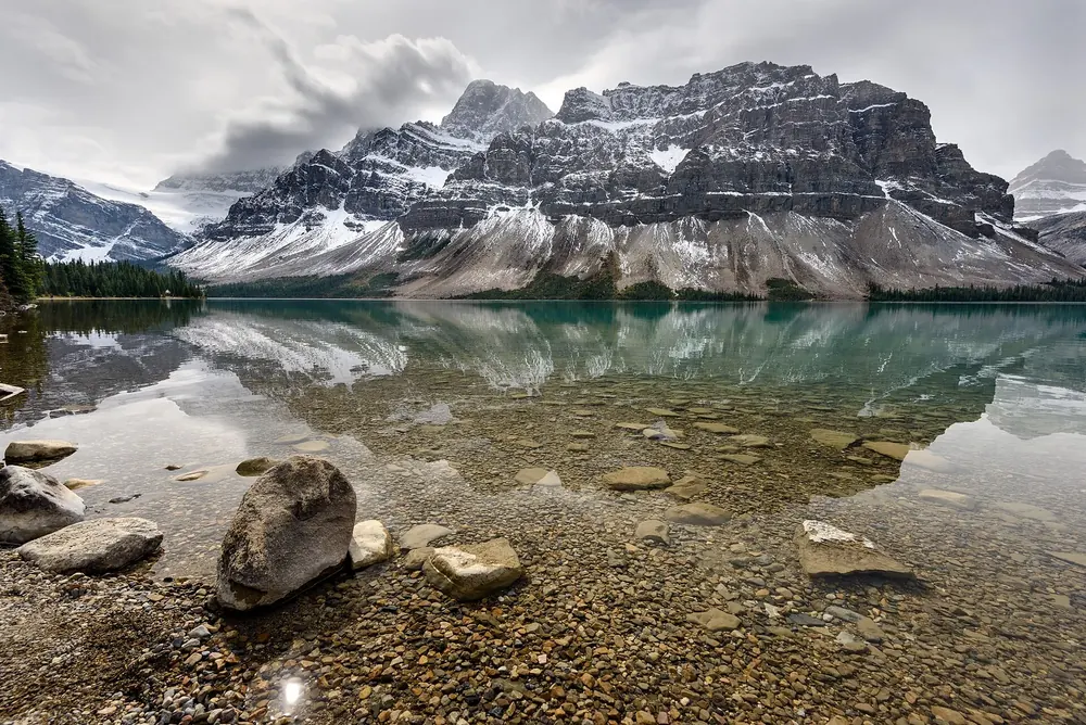 Bow Lake mit Spiegelung schneebedeckter Felsgipfel, im klaren Wasser Steine am Ufer unter Wolken