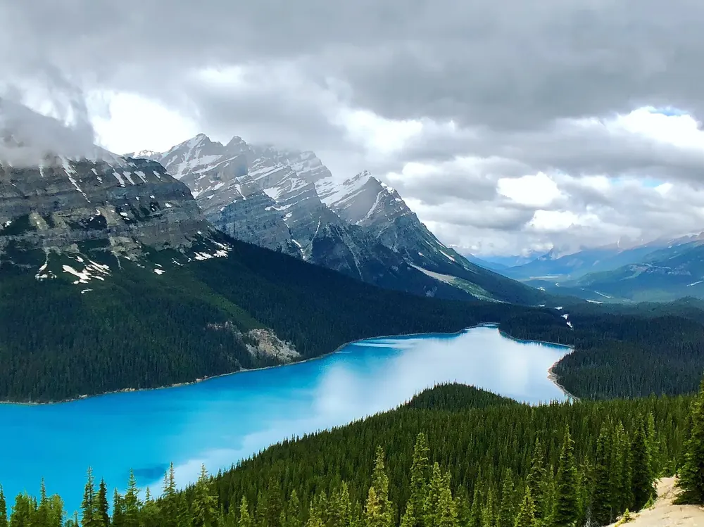 Blick vom Bow Summit auf den leuchtend türkisfarbenen Peyto Lake, bewaldetes Tal und wolkenverhangene Gipfel