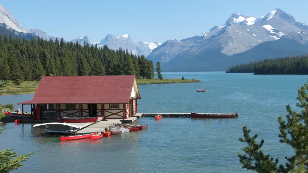 Rotes Bootshaus am Maligne Lake mit Steg und roten Kanus, dahinter Nadelwald und schneebedeckte Gipfel der Rocky Mountains