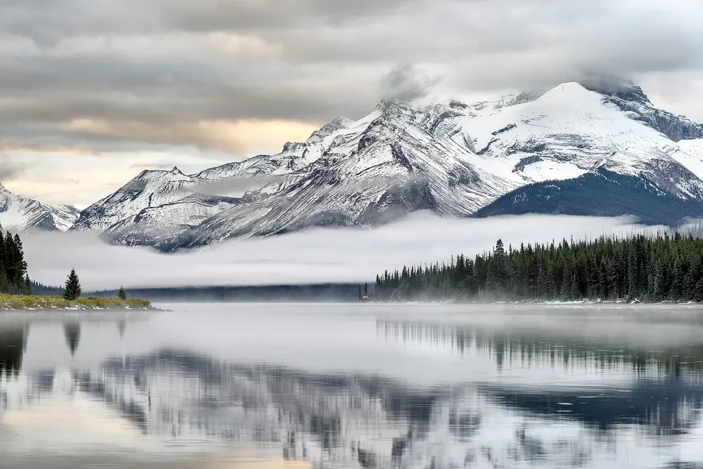 Sonnenaufgang am Maligne Lake: Nebelschwaden über dem stillen See vor verschneiten Bergen und Nadelwald