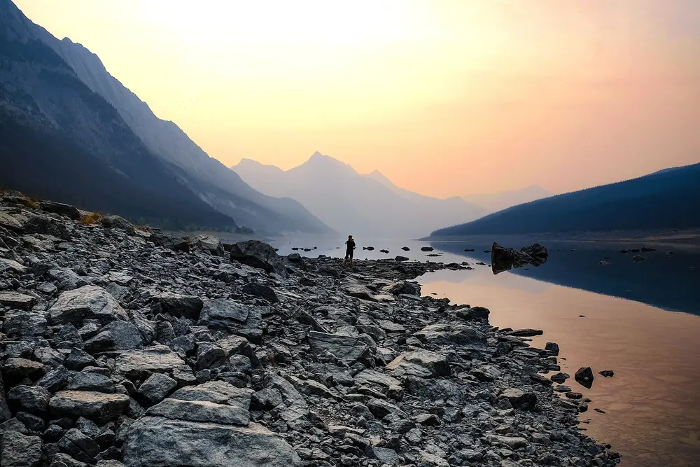 Medicine Lake im Morgenlicht: felsiges Ufer, stiller See und dunstige Bergketten im Rauch naher Waldbrände