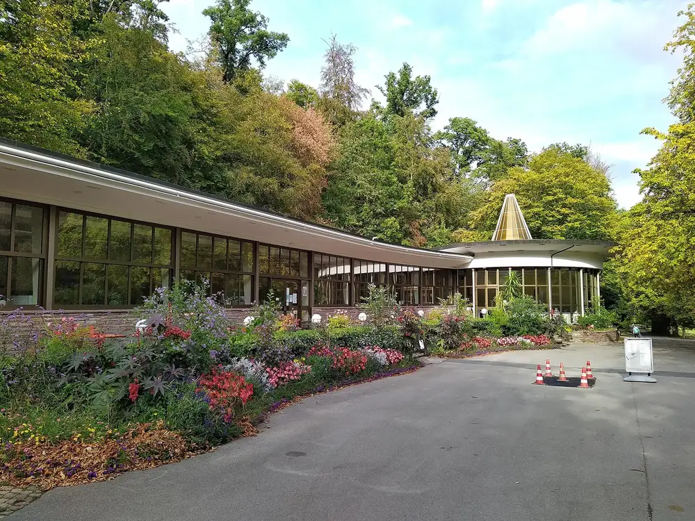 Kurpark Bad Wildungen: Wandelhalle mit Glasfront und spitzem Pavillon hinter üppigen Blumenbeeten und Bäumen