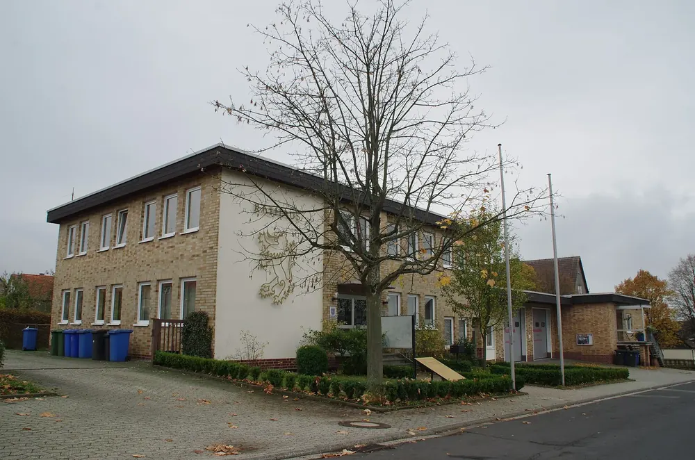 Rathaus von Frankenau: zweistöckiges Gebäude aus Naturstein und Putz mit Fahnenmasten unter kahlen Bäumen