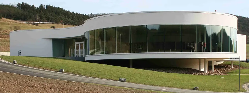NationalparkZentrum Kellerwald: moderner, geschwungener weißer Bau mit großer Glasfront zwischen Wiesen und Hügeln