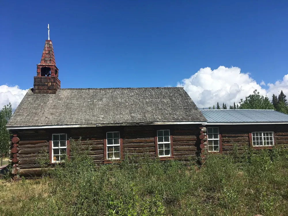 Alte Blockhauskirche in Burwash Landing mit hölzernem Glockentürmchen auf einer Wiese unter blauem Himmel