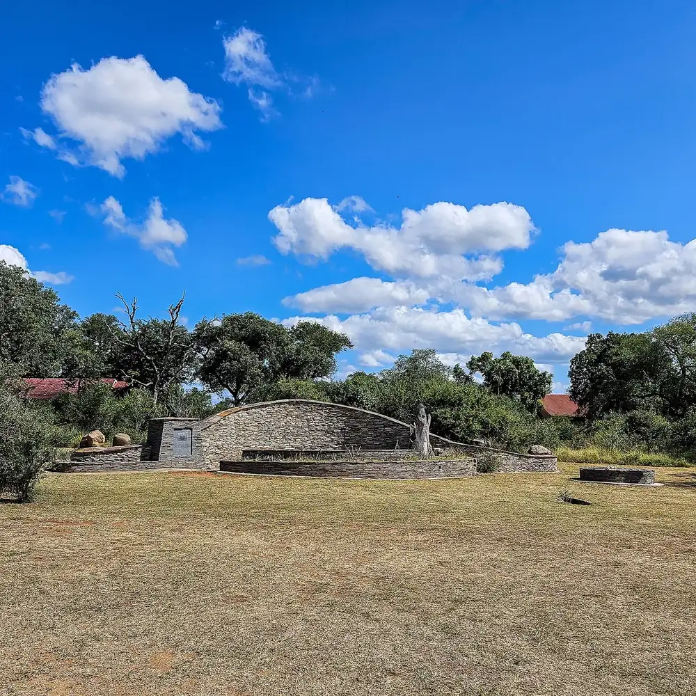 Denkmal am Paul Kruger Gate: halbrunde Natursteinmauer auf trockener Wiese zwischen Bäumen