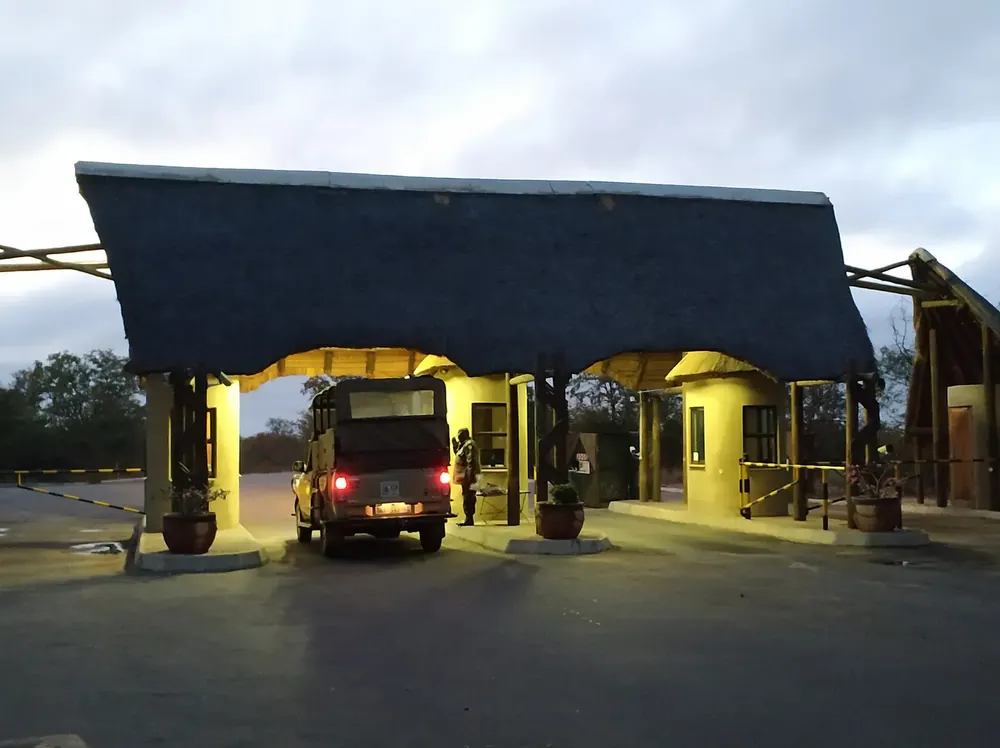 Phalaborwa Gate bei Sonnenaufgang: beleuchtetes Torgebäude mit großem Strohdach über der Zufahrt, darunter ein Safarifahrzeug