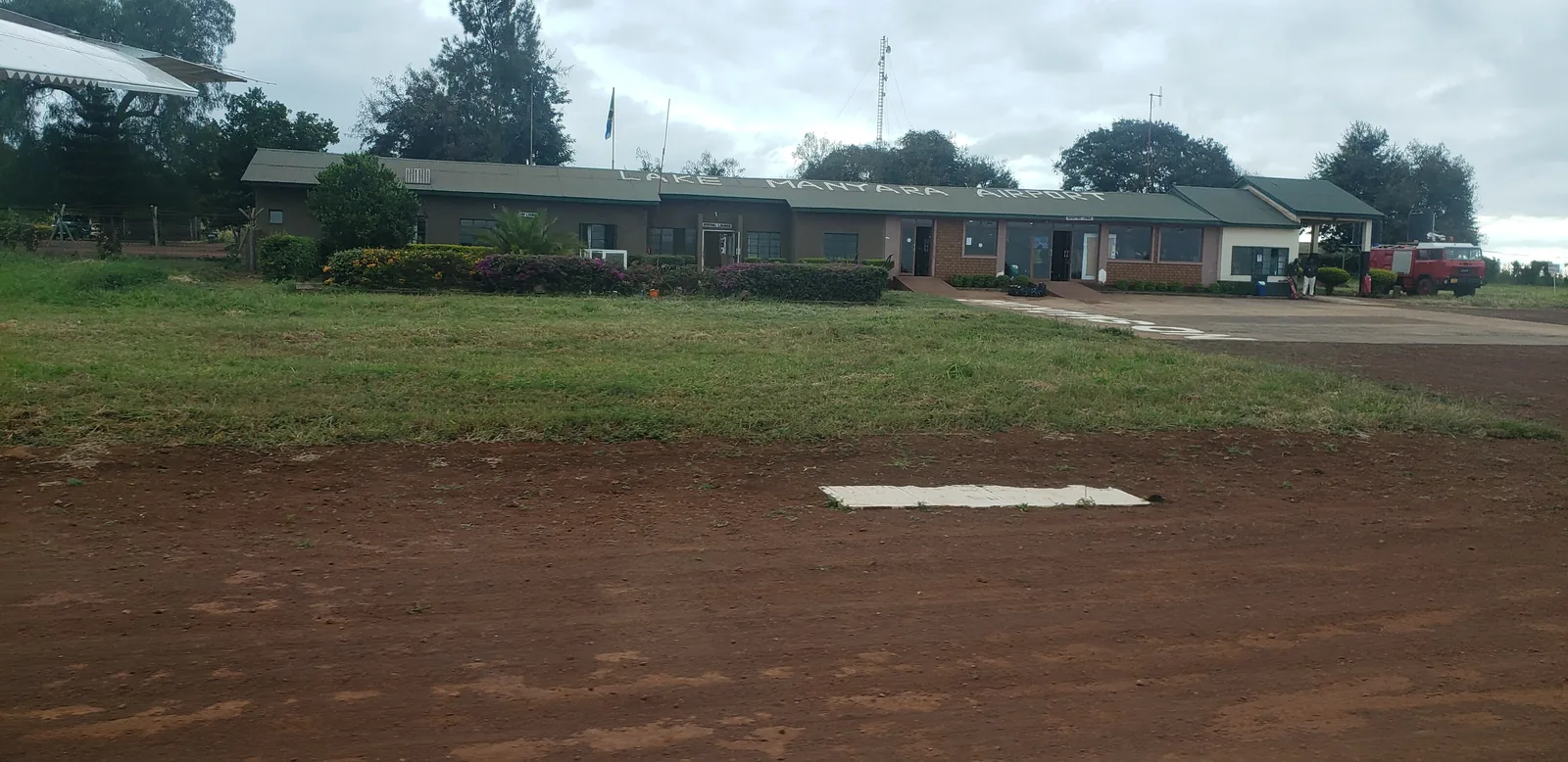 Flaches Abfertigungsgebäude des Lake Manyara Airport hinter einer Wiese und dem roten Vorfeld, außerhalb des Nationalparks