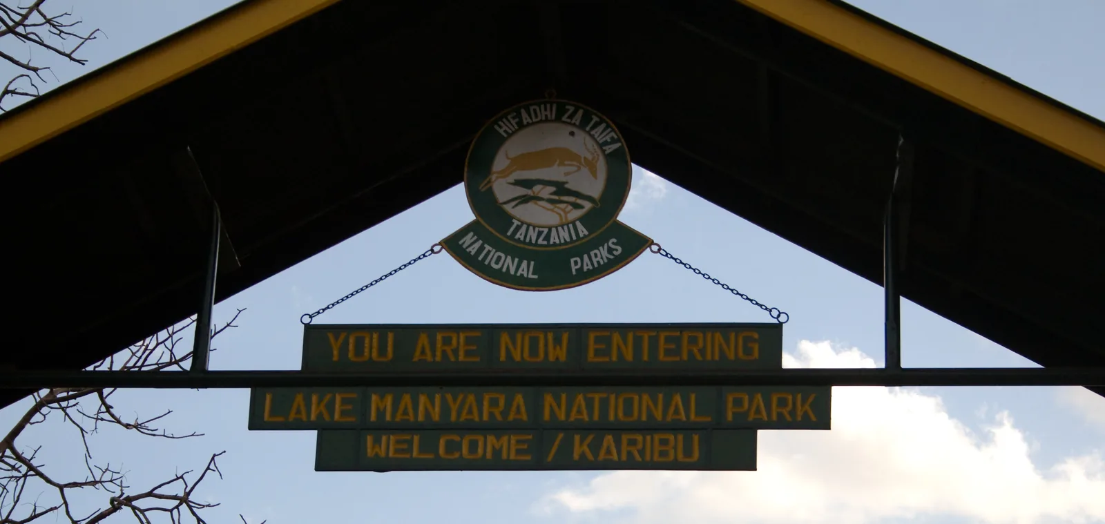 Torbogen am Haupteingang des Lake-Manyara-Nationalparks mit dem Schild „You are now entering Lake Manyara National Park – Welcome/Karibu“ und dem Parkwappen
