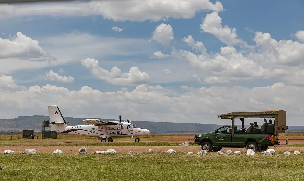 Musiara Airstrip: kleines Propellerflugzeug und ein Safari-Geländewagen auf der Graspiste unter Quellwolken