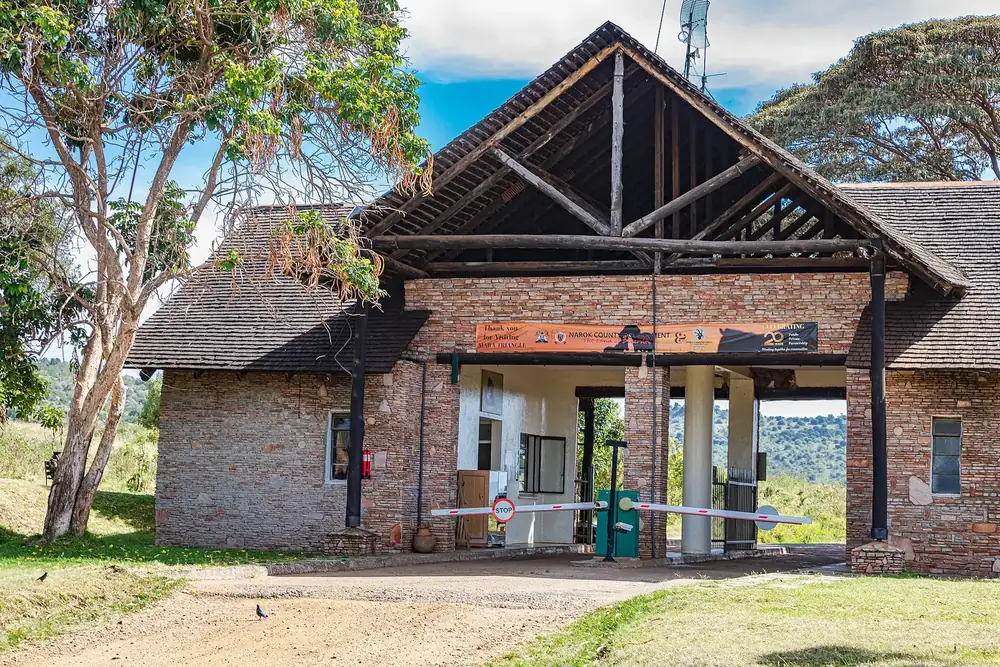 Oloololo Gate: Torgebäude aus Backstein mit hohem Holzgiebel und Schranke, daneben Akazien