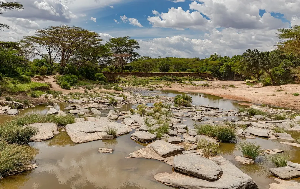 Talek River: flaches Flussbett mit Felsplatten und Wasserlachen, gesäumt von Akazien unter Quellwolken