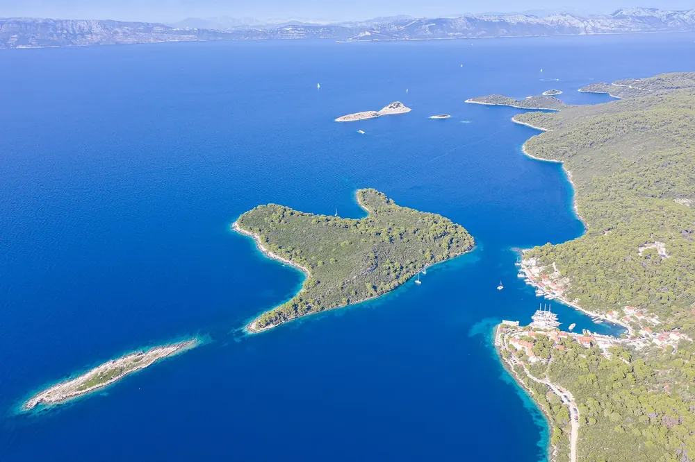 Luftbild von Pomena auf Mljet: kleiner Hafenort an einer Bucht, davor die bewaldete Insel Pomeštak im tiefblauen Meer