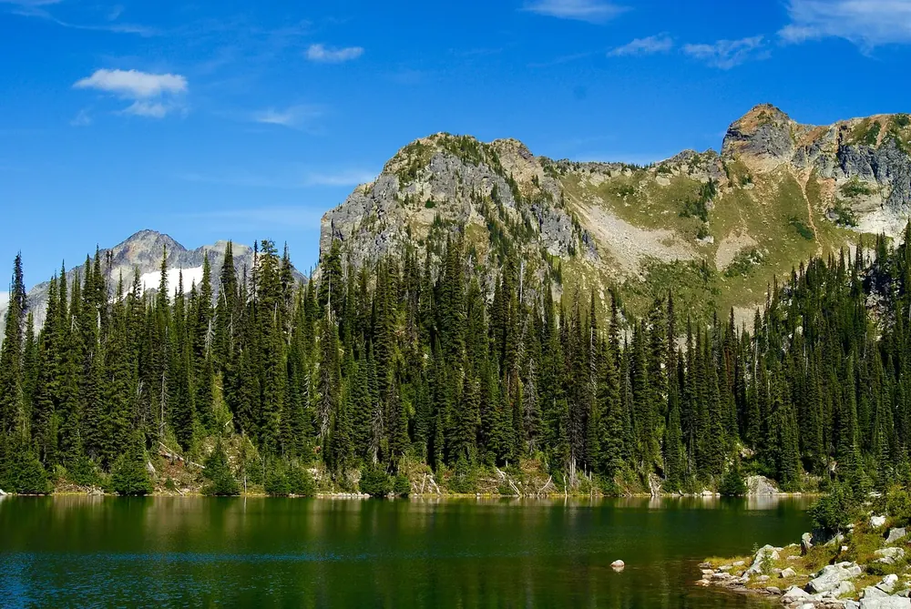 Eva Lake: tiefblauer Bergsee vor dichtem Nadelwald und felsigen Gipfeln unter blauem Himmel
