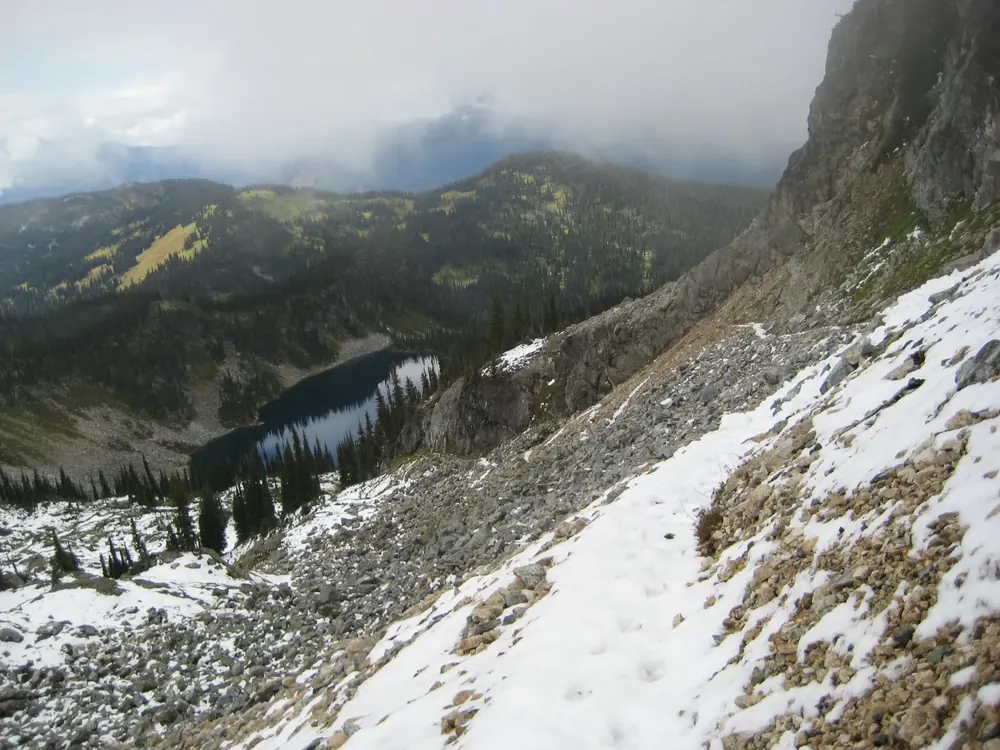 Blick vom Jade Pass Trail hinab auf den dunklen Miller Lake zwischen Schneefeldern, Geröll und Nadelwald