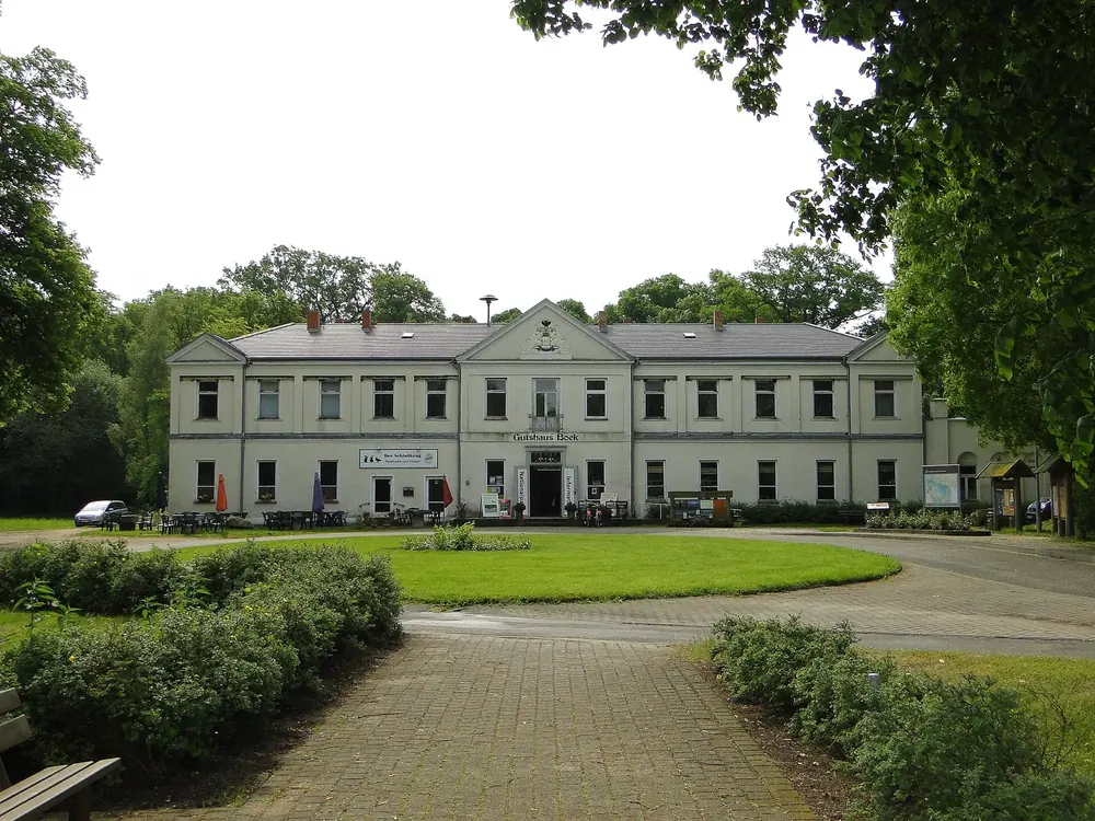 Ehemaliges Herrenhaus in Boek: langgestreckter hellgrauer Bau mit Mittelrisalit hinter einer Rasenfläche mit Hecken