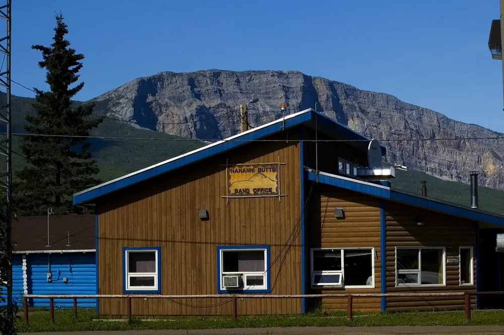 Gemeindebüro in Nahanni Butte: Holzhaus mit blauen Kanten vor der steilen Felswand des Berges Nahanni Butte