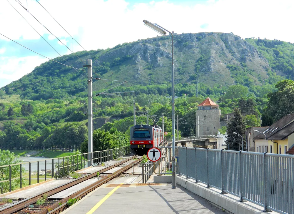 Ein Zug fährt aus dem Personenbahnhof Hainburg an der Donau, dahinter der Braunsberg am Rand des Nationalparks Donau-Auen