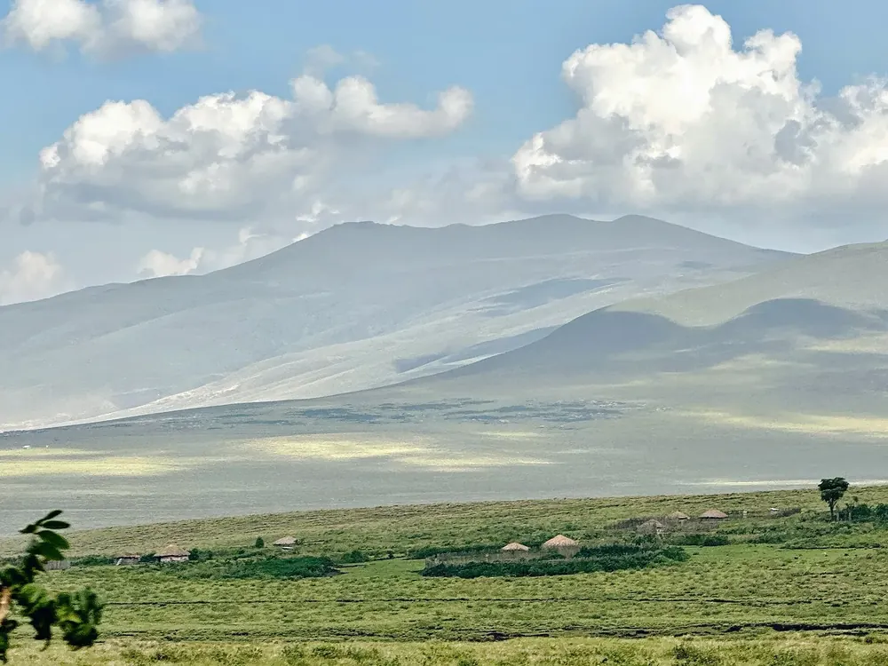 Blick von Nainokanoka über grüne Hochweiden mit Maasai-Gehöften zum Berg Loolmalasin unter Quellwolken