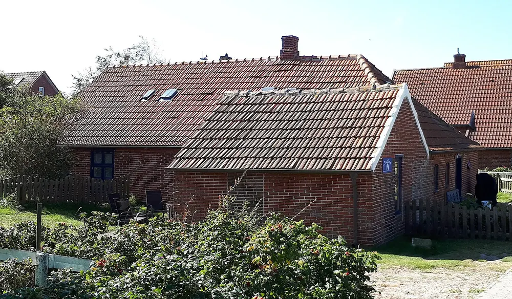Backsteinhaus mit rotem Ziegeldach im Ostdorf auf Baltrum, davor Holzzaun und Gartenbewuchs