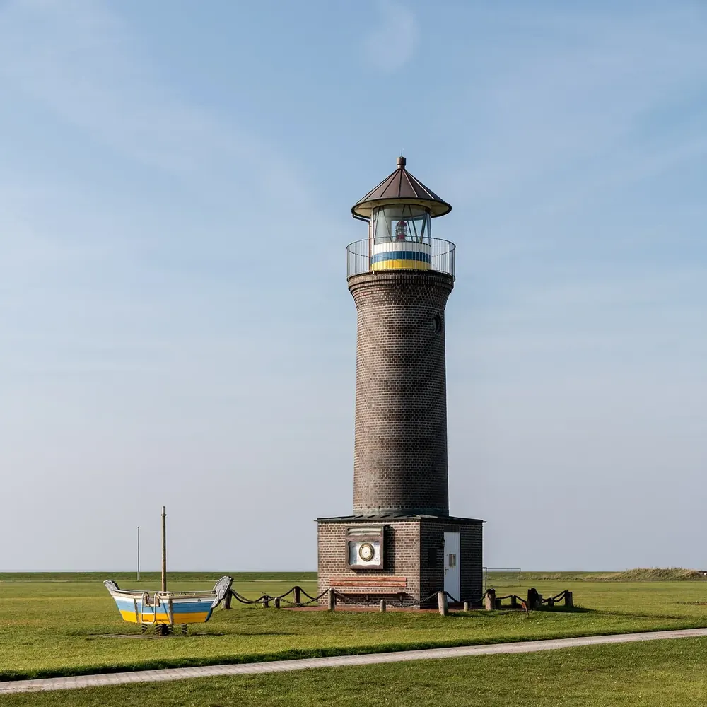 Memmertfeuer am Hafen von Juist: runder Backsteinleuchtturm auf grüner Wiese, daneben ein kleines Boot