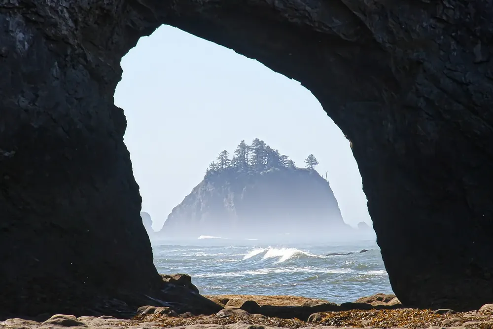 Hole-in-the-Wall: Blick durch einen dunklen Felsbogen am Strand auf eine bewaldete Felsinsel im Meeresdunst