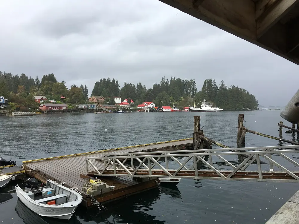 Hafen von Bamfield: Holzsteg mit Booten im Vordergrund, gegenüber Häuser am bewaldeten Ufer des Meeresarms
