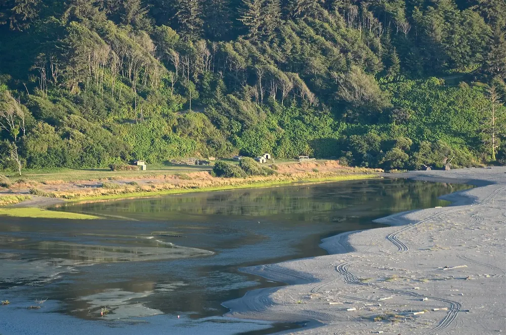 Mündung des Klamath River: ruhiges Wasser hinter einer Sandbank, am Ufer Holzhütten des Yurok-Stammes vor Waldhang
