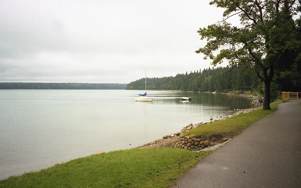 Seeufer in Wasagaming: Uferweg mit Rasen und Steinbefestigung, ein Segelboot vor bewaldetem Ufer