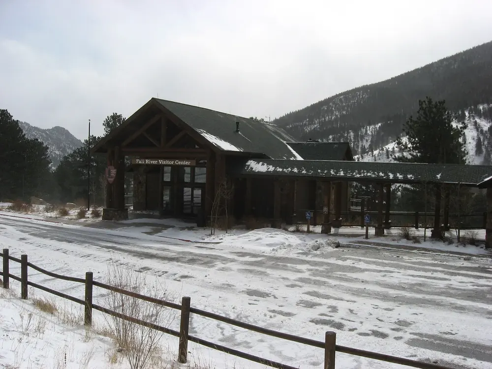 Fall River Visitor Center im Winter: Holzgebäude mit steilen Dächern, davor verschneiter Platz und Zaun, dahinter Berge