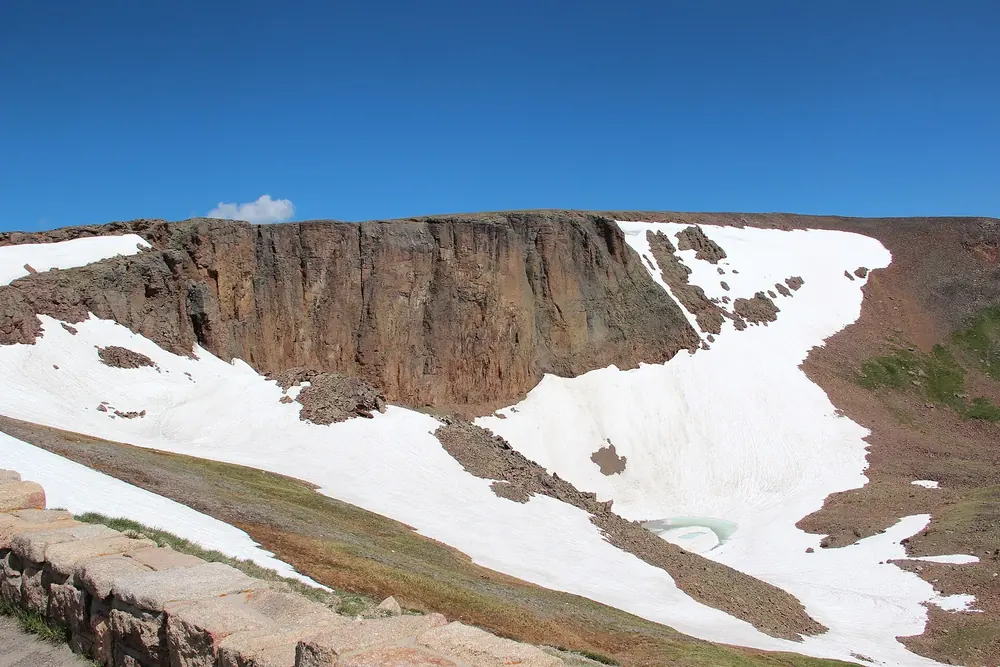 Lava Cliffs: dunkle Felswand über einem Kar mit großen Schneefeldern und Tundra unter blauem Himmel