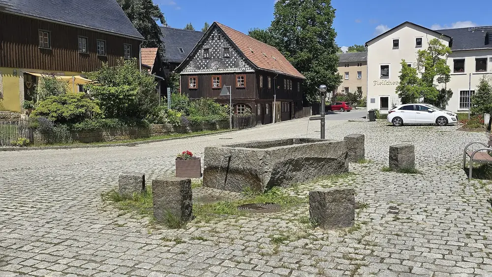 Dorfplatz von Hinterhermsdorf: Steintrog-Brunnen auf Kopfsteinpflaster, dahinter Umgebindehäuser mit Schieferfassade