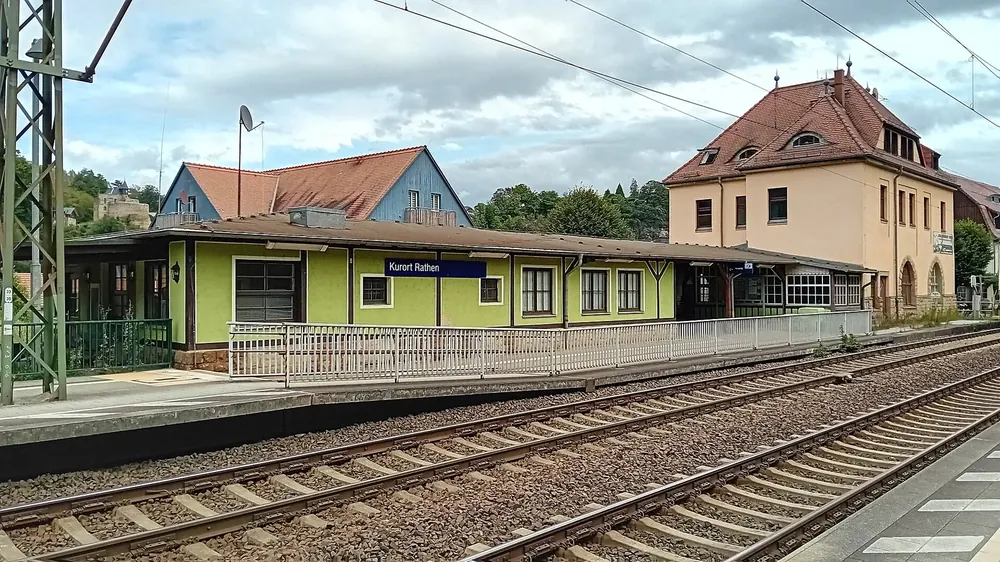 Bahnhof Kurort Rathen: grünes Bahnsteiggebäude und historisches Empfangsgebäude hinter den Gleisen der Elbtalstrecke