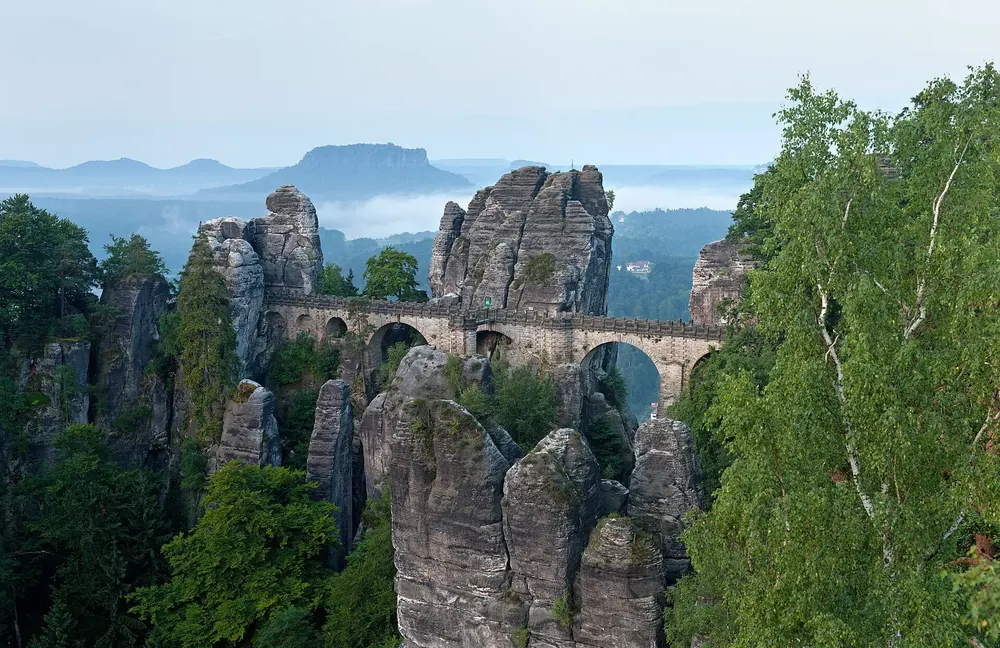 Basteibrücke am Morgen: steinerne Bogenbrücke zwischen zerklüfteten Sandsteinfelsen, dahinter Lilienstein im Dunst