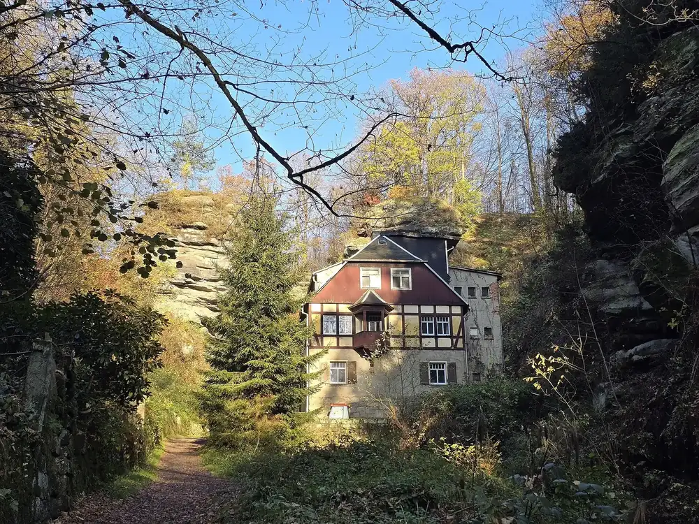 Rathewalder Mühle nahe dem Parkplatz Bastei: Fachwerkhaus zwischen Sandsteinfelsen und Nadelbäumen, davor ein Fußweg